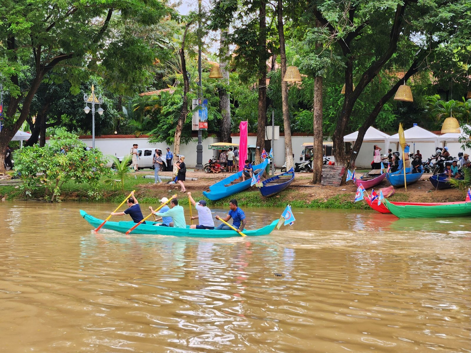A turquoise canoe team paddles past beached boats on the riverbank