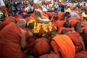 Monks in orange robes conduct a religious ceremony around a golden Buddha statue outdoors.